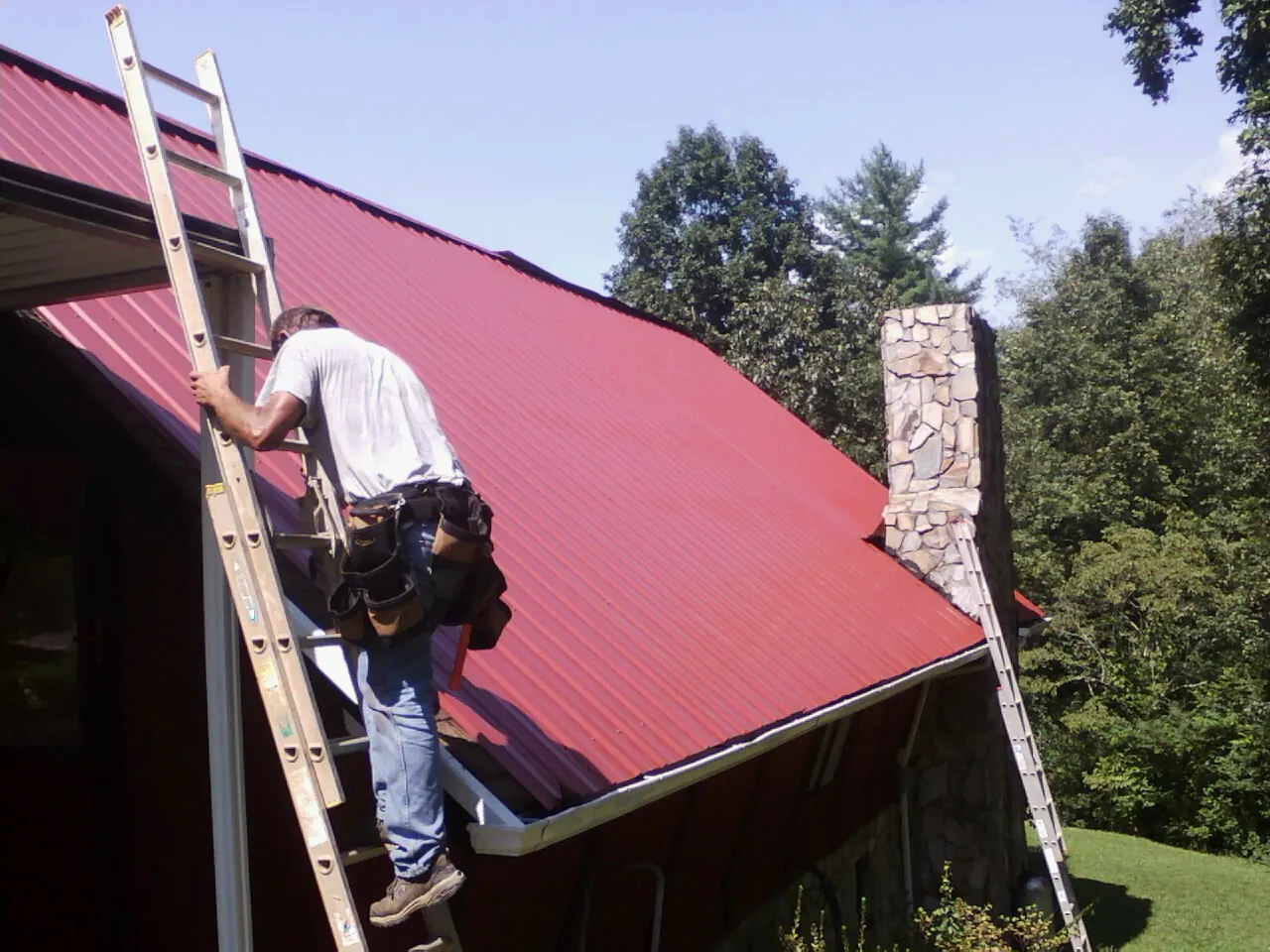 Roofer on a ladder beside a red metal roof and stone chimney — Soffit & Fascia Repair by Roofing Brothers in Medina